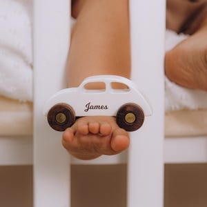 May include: A white wooden toy car with dark brown wheels and a gold-toned center detail. The side of the car features the name James printed in a black cursive script font. The toy has a smooth, rounded silhouette and a cutout window design.