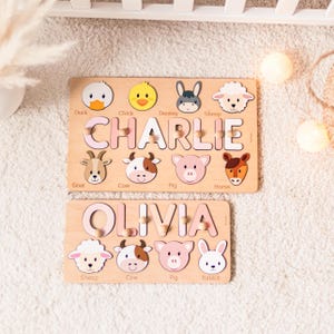 Puede incluir: Dos rompecabezas de nombres de madera con ilustraciones de animales de granja. El rompecabezas superior deletrea "CHARLIE" en letras rosas, con un pato, un pollito, un burro, una oveja, una cabra, una vaca, un cerdo y un caballo. El rompecabezas inferior deletrea "OLIVIA".