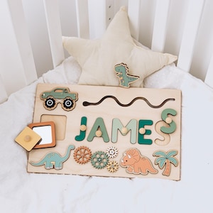 May include: A rectangular wooden activity board featuring the name JAMES in large, alternating dark and light green letters with small wooden knobs. The board includes cutouts for a green truck, a long-necked dinosaur, gears, a triceratops, and a palm tree.