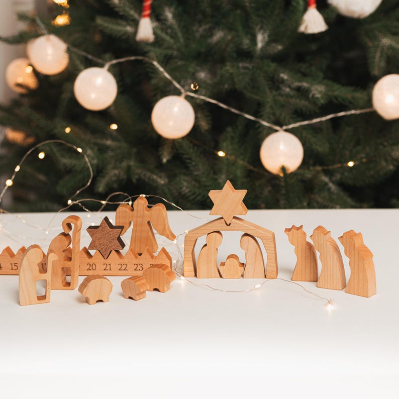 May include: Wooden nativity scene set with figures of Mary, Joseph, baby Jesus, an angel, three wise men, a shepherd, a sheep, and a star. The set is arranged on a white surface with a Christmas tree in the background.