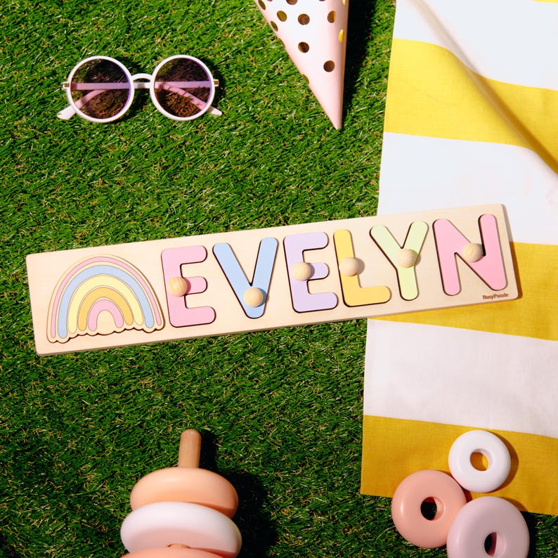 May include: A wooden puzzle spelling "EVELYN" in pastel colors, with a rainbow design. The puzzle is on a green surface with sunglasses, a party hat, and other toys. The letters have wooden pegs for easy handling.