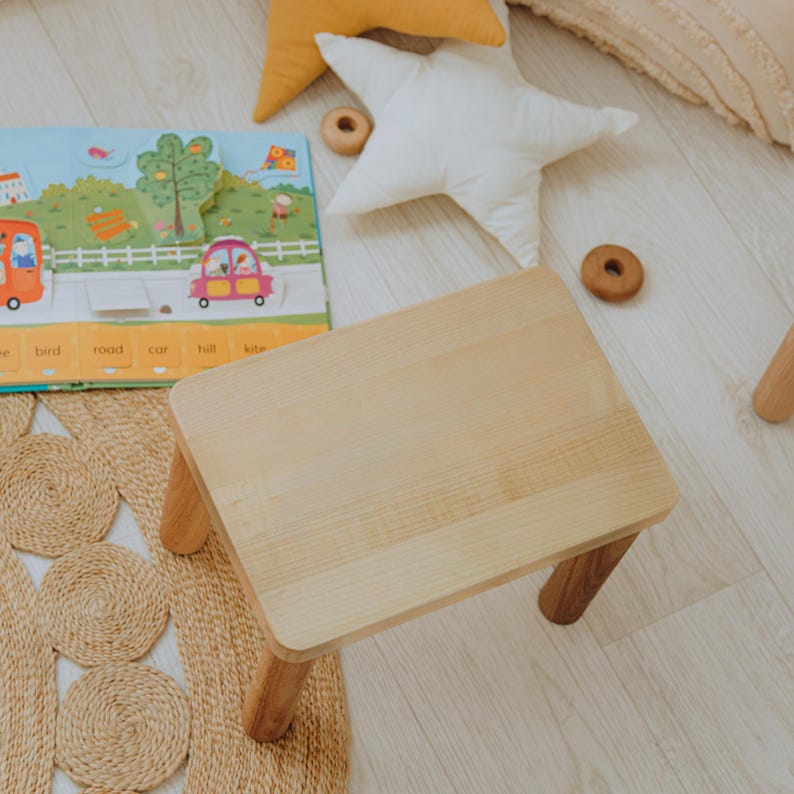 May include: A small, rectangular wooden stool with a light-colored, smooth top and four sturdy, cylindrical legs. The stool is positioned near a colorful children's book featuring illustrations of cars, trees, and a kite, alongside two star-shaped pillows.