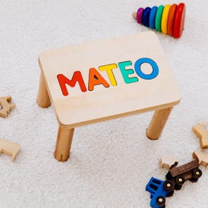 May include: A light-colored wooden stool featuring the name MATEO in large, colorful, cutout letters on the top surface. The letters are red, orange, yellow, green, and blue. The stool has four sturdy, cylindrical wooden legs and a smooth finish.