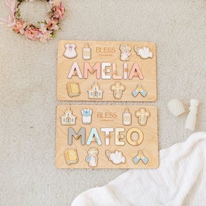 May include: Two wooden name puzzles, one reading "AMELIA" in pink and the other "MATEO" in gray, with religious-themed puzzle pieces. The word "BLESS" is printed above each name. A floral wreath and white blanket are in the background.