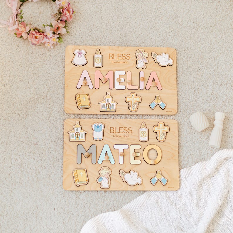 May include: Two wooden name puzzles, one reading "AMELIA" in pink and the other "MATEO" in gray, with religious-themed puzzle pieces. The word "BLESS" is printed above each name. A floral wreath and white blanket are in the background.