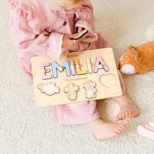 May include: Wooden name puzzle featuring the name "EMILIA" in pastel colors. The puzzle includes decorative shapes and the words "BLESS" and "Forevermore". A child is holding the puzzle. The puzzle is made of wood.