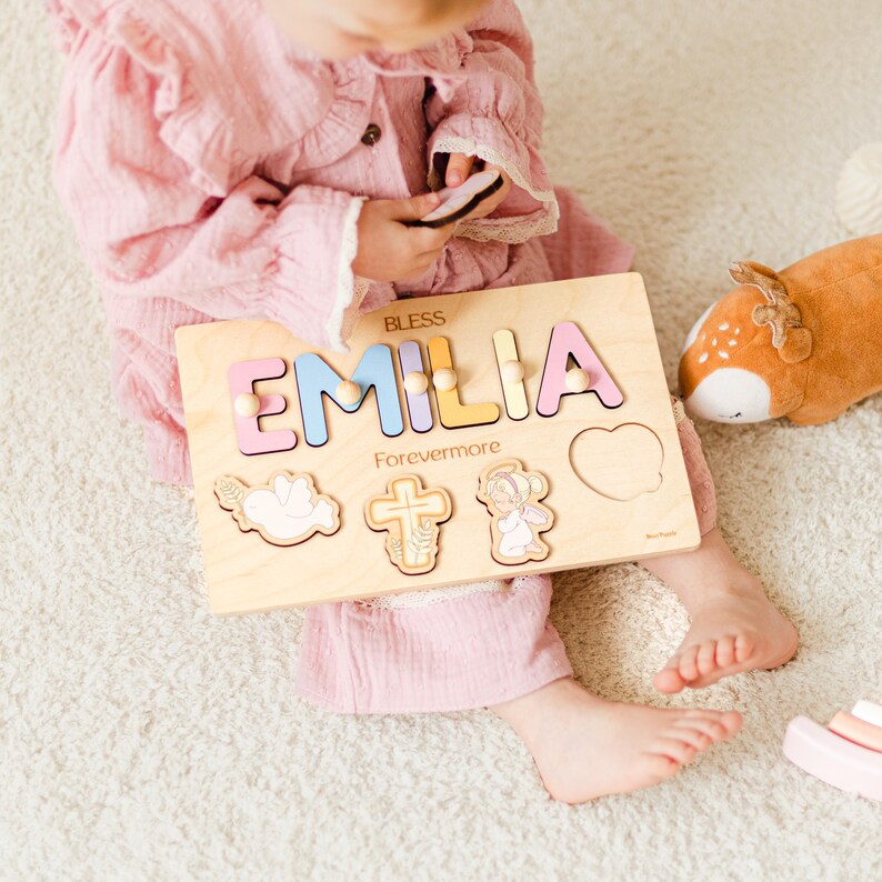 May include: Wooden name puzzle featuring the name "EMILIA" in pastel colors. The puzzle includes decorative shapes and the words "BLESS" and "Forevermore". A child is holding the puzzle. The puzzle is made of wood.
