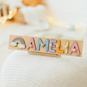 May include: Wooden name puzzle spelling "AMELIA" in pastel colors, with a rainbow and clouds design. Each letter has a wooden peg for easy handling. The puzzle is displayed on a white surface.