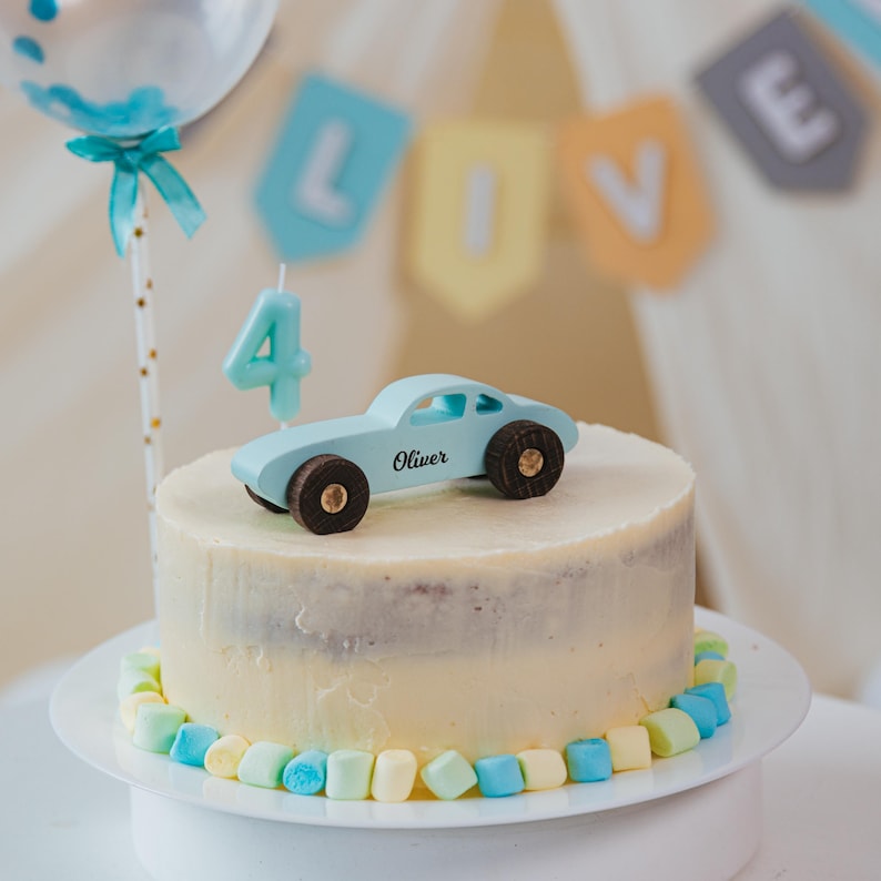 Puede incluir: Un pastel redondo con glaseado blanco, decorado con un borde de malvaviscos azul claro y amarillo p&aacute;lido. Un coche de juguete azul claro con el nombre Oliver impreso en el lateral y una vela azul claro con el n&uacute;mero cuatro sobre el pastel.