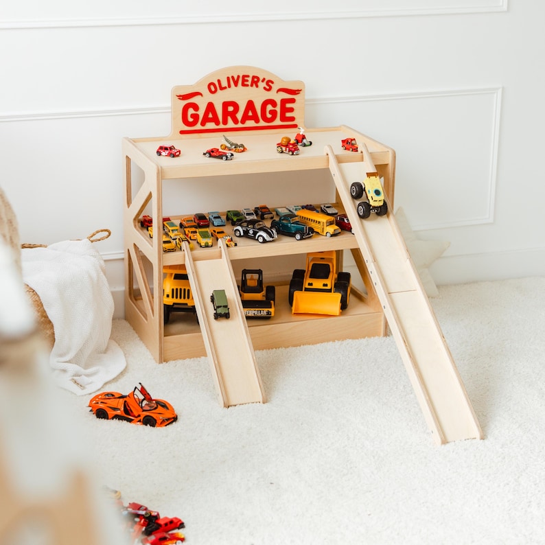 May include: A light-colored wooden toy garage with two levels, two ramps, and a sign at the top that reads OLIVER'S GARAGE in red letters. The garage is filled with various small toy cars, trucks, and construction vehicles in multiple colors.