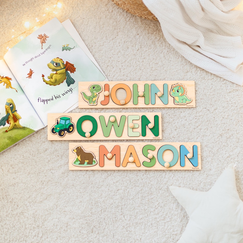 May include: Three wooden name puzzles with the names "JOHN", "OWEN", and "MASON" in colorful letters. Each puzzle features a different animal or vehicle: a dinosaur, a tractor, and a bear. A children's book is open in the background.
