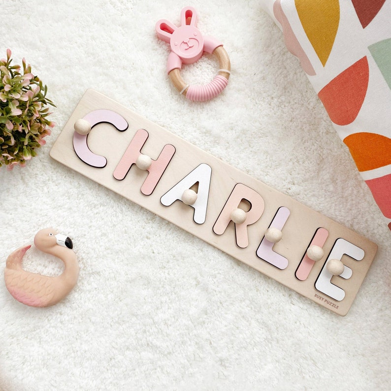 Custom name puzzle with pegs at extra charge. 1st birthday personalized gift. montessori toddler toys.Pink and white colors letters, wooden base. Their length varies from 8 to 24 in., depending on the number of letters. The puzzle width is 3,35 in.