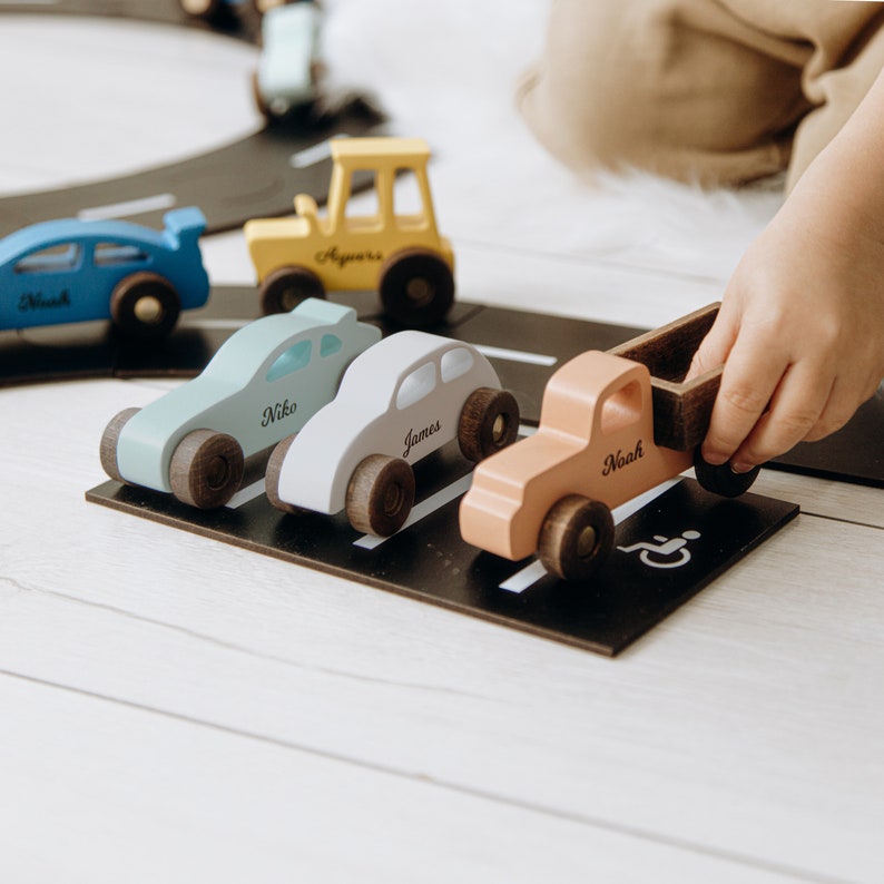 Puede incluir: Una colecci&oacute;n de peque&ntilde;os coches de juguete de madera en azul, amarillo, azul claro, blanco y melocot&oacute;n, cada uno con un nombre impreso en el lateral. Los veh&iacute;culos est&aacute;n dispuestos sobre una alfombrilla de aparcamiento negra con un s&iacute;mbolo blanco.