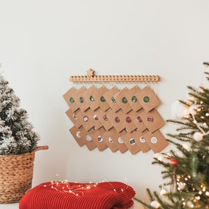 Op de afbeelding: Een adventkalender van bruin papier met 24 genummerde enveloppen die aan een houten balk hangen. Elke envelop heeft een kleine, kleurrijke sticker. De kalender hangt aan een witte muur met een kerstboom en lichtjes op de achtergrond.