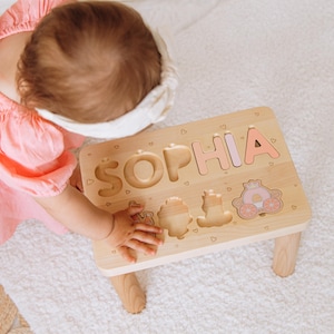 May include: A light-colored wooden puzzle table featuring the name SOPHIA in large, cutout letters. The puzzle includes a pink carriage-shaped piece and small engraved heart patterns across the surface, supported by four sturdy wooden legs.