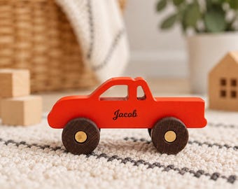 May include: A bright red wooden toy truck featuring a cutout window design and dark brown wheels with gold-toned centers. The side of the truck is personalized with the name Jacob printed in a black cursive script font on the body of the vehicle.