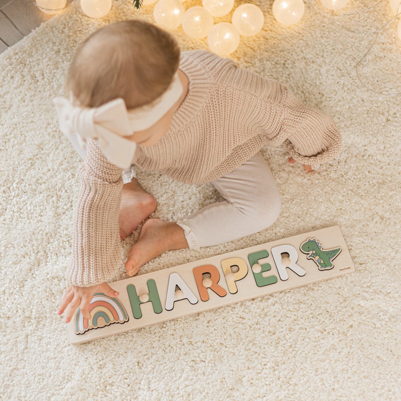 Personalized wooden name puzzle “Harper” with rainbow and dinosaur elements used by a toddler in a cozy, softly lit nursery setting. Montessori toy with chunky letters and wooden knobs for easy grip, supporting fine motor skills, hand-eye coordination, and early name recognition. Warm neutral decor with soft rug and lights. Perfect custom gift for baby, first birthday, or baby shower. Handmade wooden educational toy.
