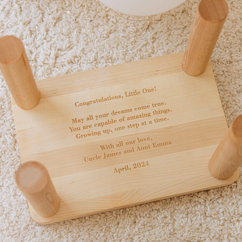May include: A light-colored wooden step stool with four cylindrical legs. The top surface features laser-engraved text: Congratulations, Little One! May all your dreams come true. You are capable of amazing things. Growing up, one step at a time. With all our love, Uncle James and Aunt Emma. April, 2024.