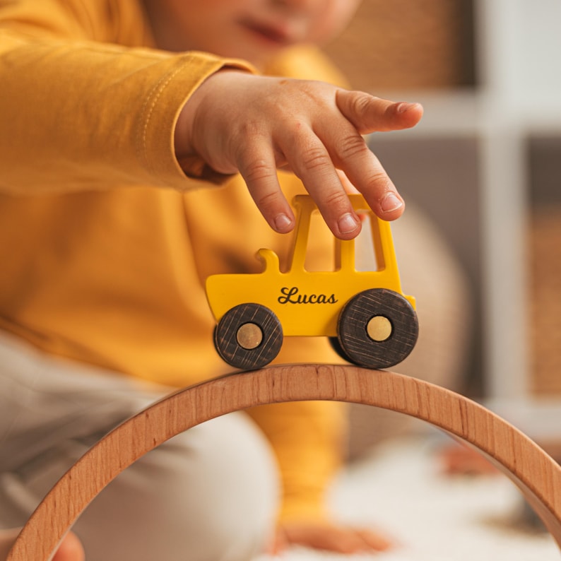 K&ouml;nnte beinhalten: Ein leuchtend gelber Spielzeugtraktor aus Holz mit dem Namen Lucas in schwarzer Schreibschrift auf der Seite. Der Traktor hat vier dunkelbraune R&auml;der mit sichtbarer Holzmaserung und steht auf einem gebogenen Holzbogenteil.