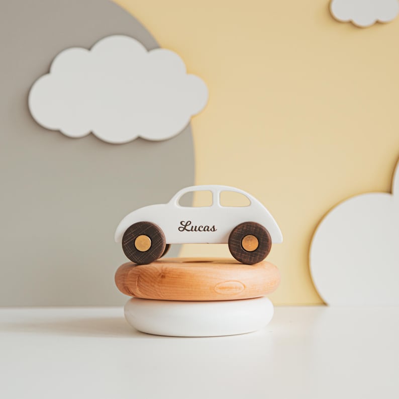 Puede incluir: Un coche de juguete de madera blanca con ruedas de color marr&oacute;n oscuro y centros dorados, con el nombre Lucas impreso en cursiva negra en el lateral. El coche est&aacute; equilibrado sobre dos anillos de madera lisos y redondeados, uno natural y otro blanco.