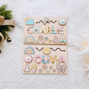 May include: Two rectangular wooden name puzzles featuring the names CHARLIE and OLIVIA in pastel-colored letters. Each board includes removable wooden shapes such as rainbows, flowers, a unicorn, a butterfly, a sun, and a hot air balloon.