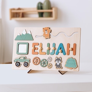 May include: A rectangular wooden activity board featuring the name ELIJAH in colorful letters, a sliding bear figure, a mirror, a green truck, rotating gears, a wolf illustration, and a pine tree shape, all mounted on a small wooden stand.