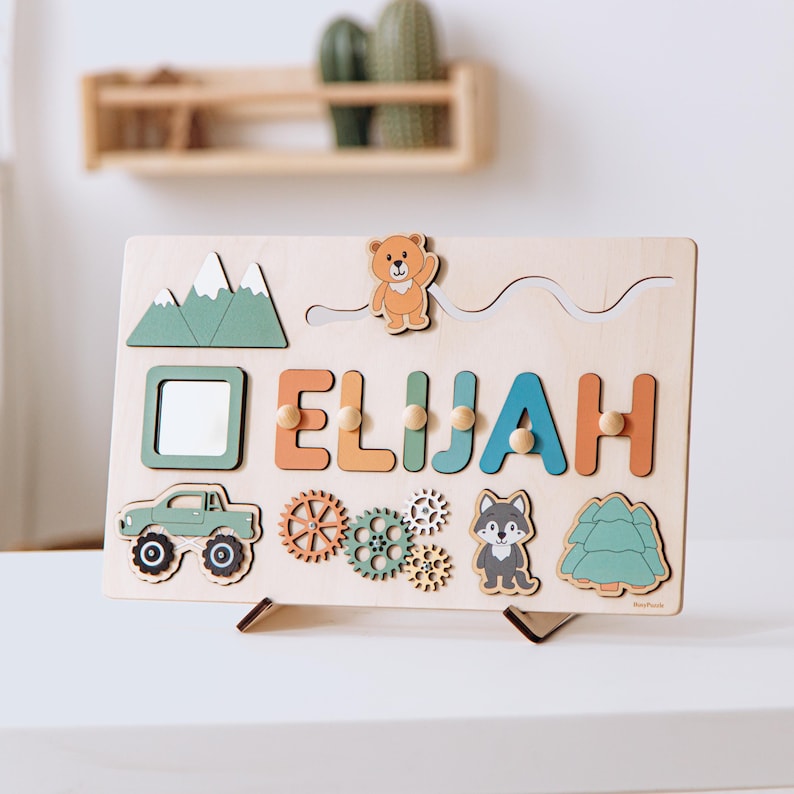 May include: A rectangular wooden activity board featuring the name ELIJAH in colorful letters, a sliding bear figure, a mirror, a green truck, rotating gears, a wolf illustration, and a pine tree shape, all mounted on a small wooden stand.