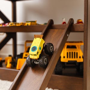 May include: A yellow monster truck toy with a porous texture and a blue circular window on the side, positioned on a wooden ramp. The ramp is part of a multi-level wooden structure containing various yellow construction vehicle toys in the background.