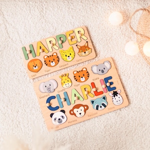 Puede incluir: Dos rompecabezas de nombres de madera, uno con el nombre "HARPER" y el otro "CHARLIE". Cada rompecabezas presenta letras coloridas y piezas de rompecabezas con temas de animales, incluyendo un le&oacute;n, un tigre y un koala. Los rompecabezas est&aacute;n sobre una superficie blanca y suave.