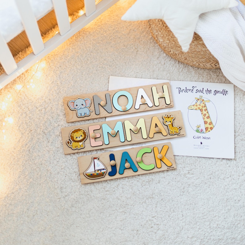 Pode incluir: Quebra-cabe&ccedil;as de madeira com os nomes "Noah", "Emma" e "Jack" em letras coloridas. Cada quebra-cabe&ccedil;a inclui uma ilustra&ccedil;&atilde;o de animal: um elefante, um le&atilde;o e uma girafa. Um livro infantil com uma girafa tamb&eacute;m &eacute; vis&iacute;vel.