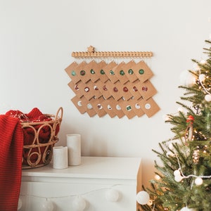 May include: A brown paper advent calendar with numbered envelopes hanging from a wooden bar. The calendar is decorated with small red and green circles. A white dresser with candles and a red blanket are in the foreground. A Christmas tree with white lights is in the background.
