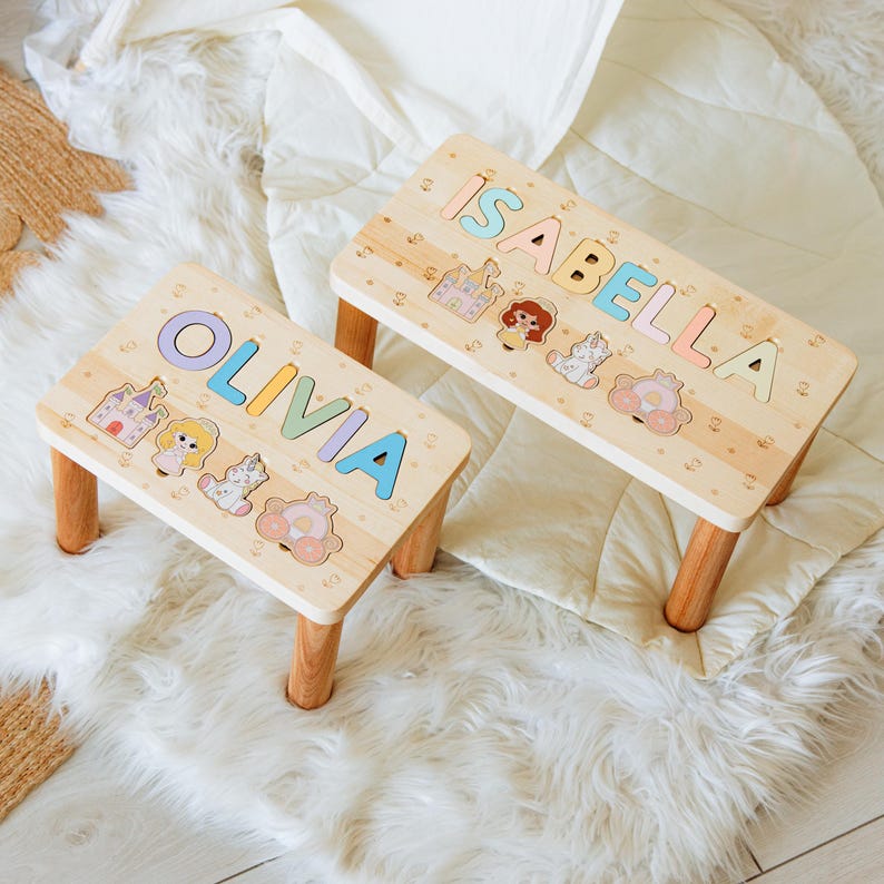 May include: Two light-colored wooden step stools with four legs each. The smaller stool features the name OLIVIA in pastel letters, while the larger stool displays the name ISABELLA. Both stools include cutouts of a castle, a girl, and a carriage.