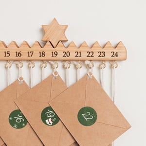 Pu&ograve; includere: Una barra per calendario dell'avvento in legno chiaro con una stella intagliata e i numeri dal 15 al 24. Piccoli ganci in metallo dorato pendono dal basso, sostenendo buste di carta marrone con cordini bianchi, sigillate da adesivi circolari verdi.