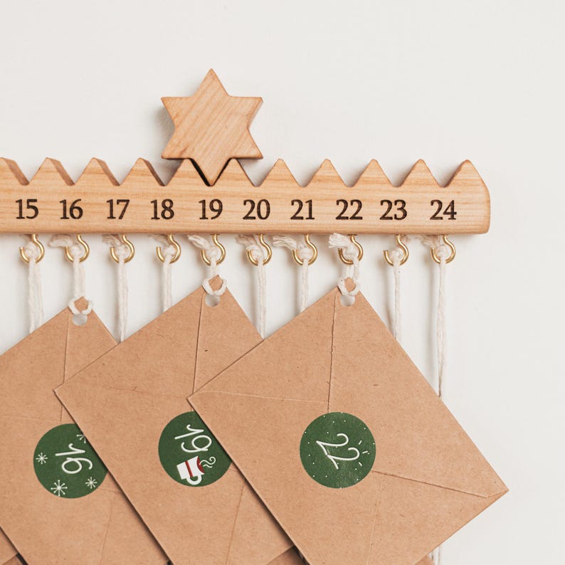 Pu&ograve; includere: Una barra per calendario dell'avvento in legno chiaro con una stella intagliata e i numeri dal 15 al 24. Piccoli ganci in metallo dorato pendono dal basso, sostenendo buste di carta marrone con cordini bianchi, sigillate da adesivi circolari verdi.