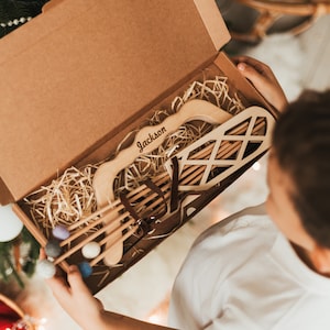 Könnte beinhalten: Ein Holzspielzeug-Bogen- und Pfeilset in einer braunen Pappbox mit dem Namen "Jackson" auf dem Bogen eingraviert. Das Set enthält einen Bogen, Pfeile und einen Köcher. Die Box ist mit braunem Verpackungsmaterial gefüllt.