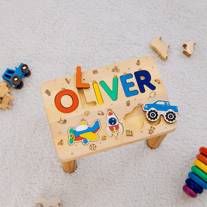 May include: A light-colored wooden puzzle board featuring the name OLIVER in colorful raised letters. The board includes cutouts for a blue truck, a red and yellow airplane, and a rocket ship, with engraved icons of various toys and vehicles.