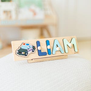 May include: Wooden name puzzle featuring the name "LIAM" in blue and green letters with wooden pegs. A police car graphic is on the left. The puzzle is on a wooden base.