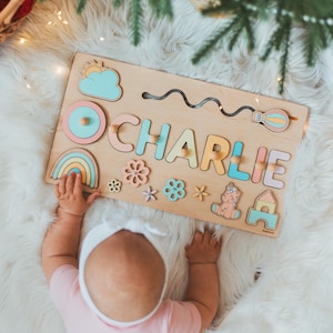 May include: A rectangular wooden name puzzle featuring the name CHARLIE in pastel-colored letters with small wooden knobs. The board includes decorative cutouts of a sun, cloud, rainbow, hot air balloon, flowers, a unicorn, and a small house.