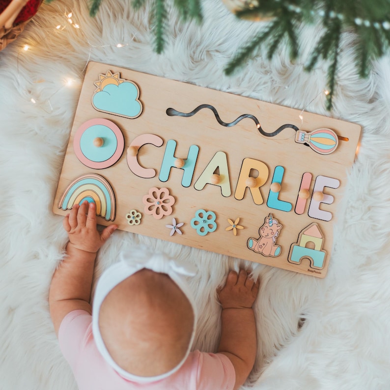 May include: A rectangular wooden name puzzle featuring the name CHARLIE in pastel-colored letters with small wooden knobs. The board includes decorative cutouts of a sun, cloud, rainbow, hot air balloon, flowers, a unicorn, and a small house.