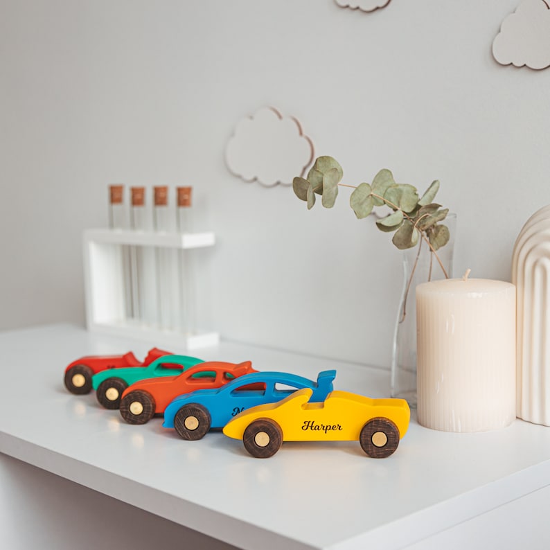 Puede incluir: Una fila de cinco coches de juguete de madera en colores rojo, turquesa, naranja, azul y amarillo. Cada coche tiene ruedas de color marr&oacute;n oscuro con centros dorados. El coche amarillo tiene el nombre Harper impreso en negro en el lateral.