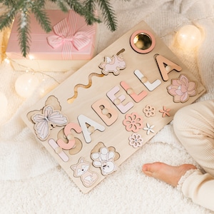May include: A wooden name puzzle board featuring the name ISABELLA in pastel pink and white letters. The board includes cutouts of a butterfly, a unicorn, a fox, a teddy bear, and various flowers, along with a circular mirror insert.