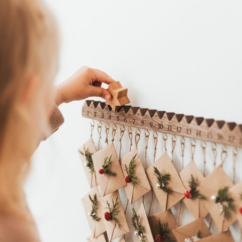 May include: A wooden advent calendar with numbered doors, each with a small brown paper envelope hanging from it. The calendar is decorated with sprigs of greenery and red berries. A hand is placing a wooden star on the calendar.