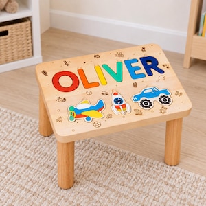 May include: A light-colored wooden step stool featuring the name OLIVER in large, multi-colored block letters. Below the name are three wooden puzzle-style cutouts of a multi-colored airplane, a rocket ship, and a blue monster truck.