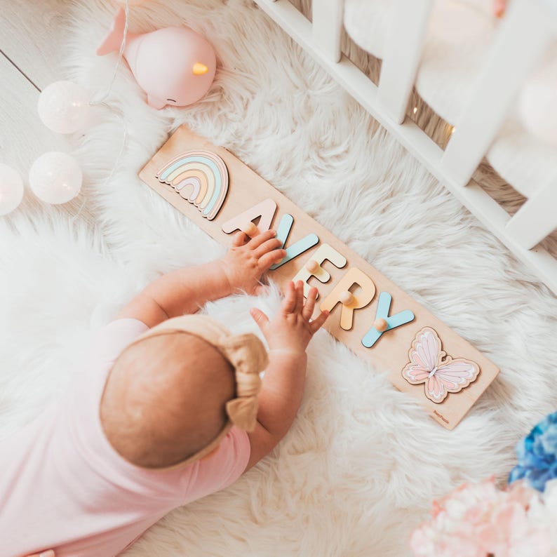 Pode incluir: Um tabuleiro de quebra-cabe&ccedil;a de madeira com o nome AVERY em letras de cores pastel, com um &iacute;cone de arco-&iacute;ris &agrave; esquerda e uma borboleta &agrave; direita. Cada letra inclui um pequeno bot&atilde;o de madeira para segurar, sobre uma base de madeira clara.