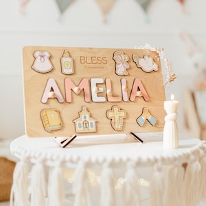 May include: Wooden puzzle board with the name "AMELIA" in pink and white letters. The board features religious-themed cutouts, including a dress, a bottle, an angel, a dove, a book, a church, a cross, and praying hands. The word "BLESS Forevermore" is also on the board.