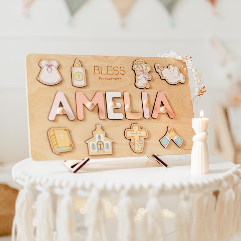 May include: Wooden puzzle board with the name "AMELIA" in pink and white letters. The board features religious-themed cutouts, including a dress, a bottle, an angel, a dove, a book, a church, a cross, and praying hands. The word "BLESS Forevermore" is also on the board.