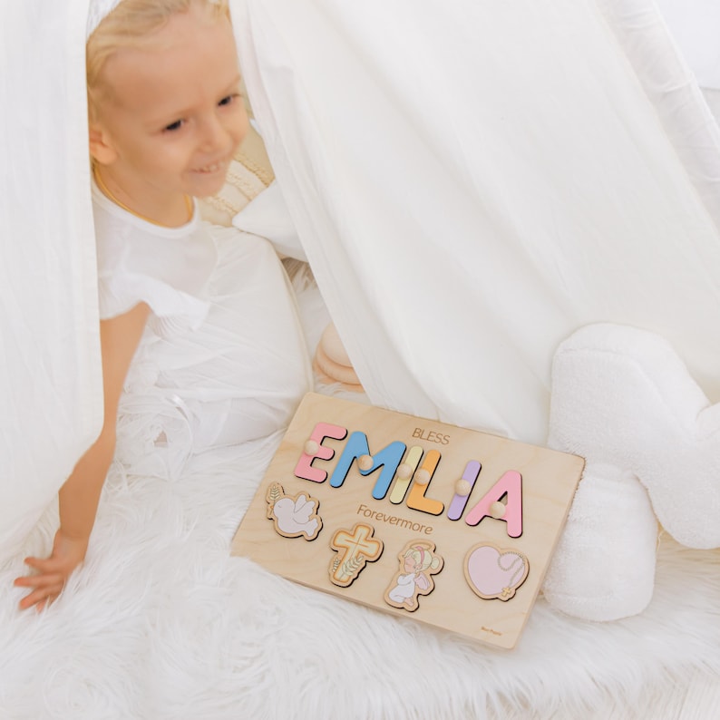 May include: Wooden puzzle with the name "EMILIA" in pastel colors. The puzzle includes the word "BLESS" and "Forevermore". The puzzle also features angel, cross, and heart shapes. The puzzle is on a white fluffy surface.