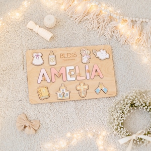 May include: Wooden puzzle with the name "AMELIA" in pink letters. The puzzle includes religious-themed shapes and the text "BLESS Forevermore". The puzzle is on a light-colored surface with decorative elements.