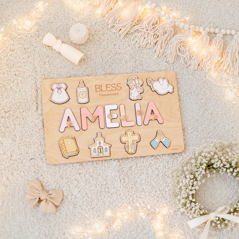 May include: Wooden puzzle with the name "AMELIA" in pink letters. The puzzle includes religious-themed shapes and the text "BLESS Forevermore". The puzzle is on a light-colored surface with decorative elements.