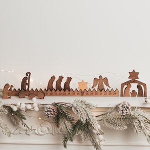 Op de afbeelding: Houten adventskalender met een kerststal en de nummers 9 tot en met 24. De kalender is versierd met een ster en een houten engel. De kalender staat op een wit oppervlak met een krans van groen.